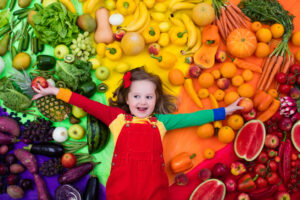 A little girl laying on the ground surrounded by fruits and vegetables.